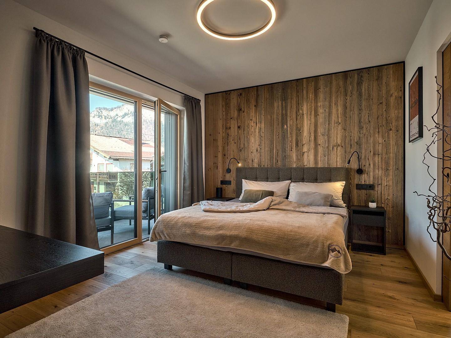 Helles Schlafzimmer mit Doppelbett, Holzwand und direktem Zugang zum Balkon mit Bergblick.
