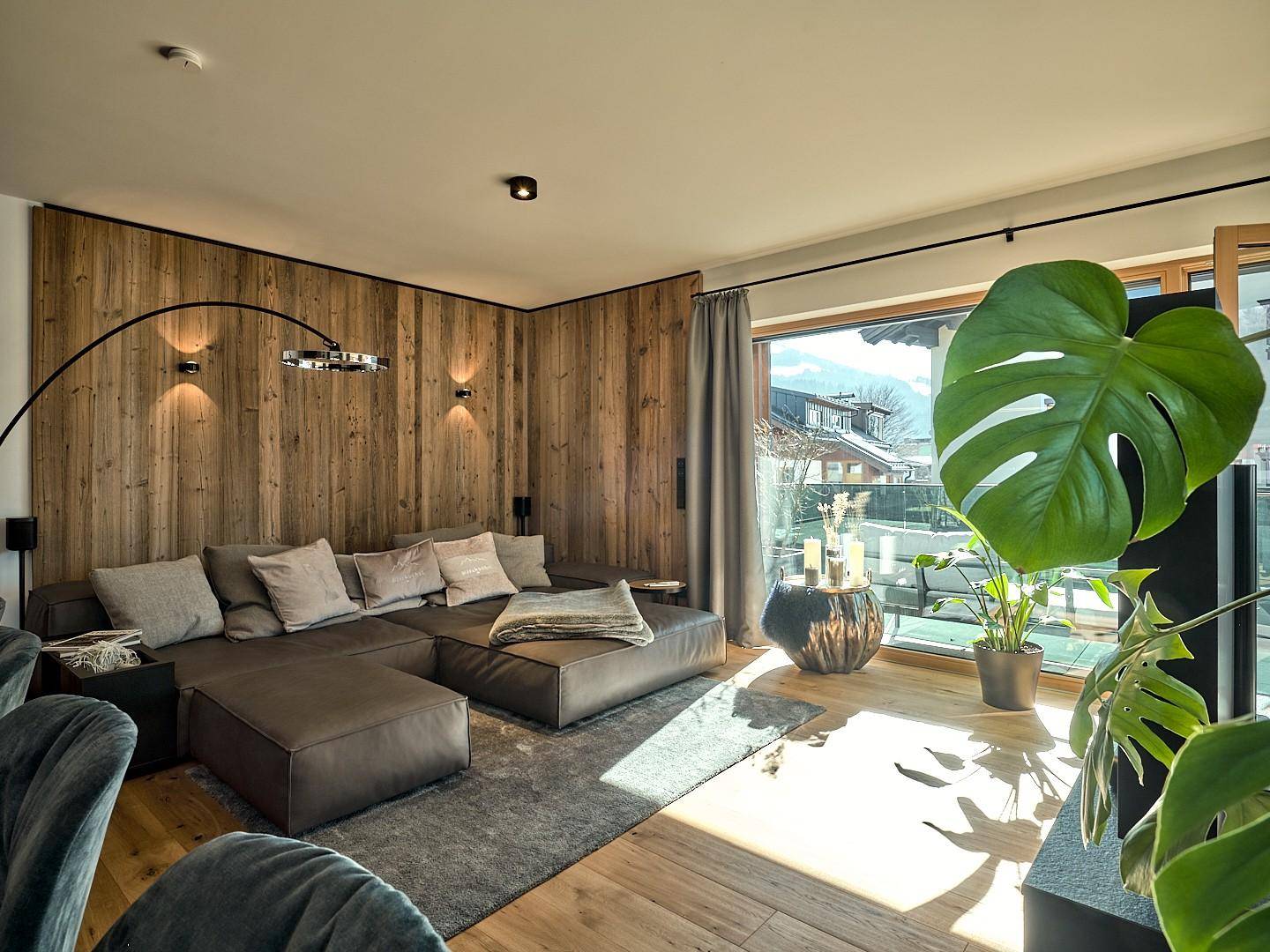 Gemütliches Wohnzimmer mit großem Sofa, Holzwand und Zugang zum Balkon.