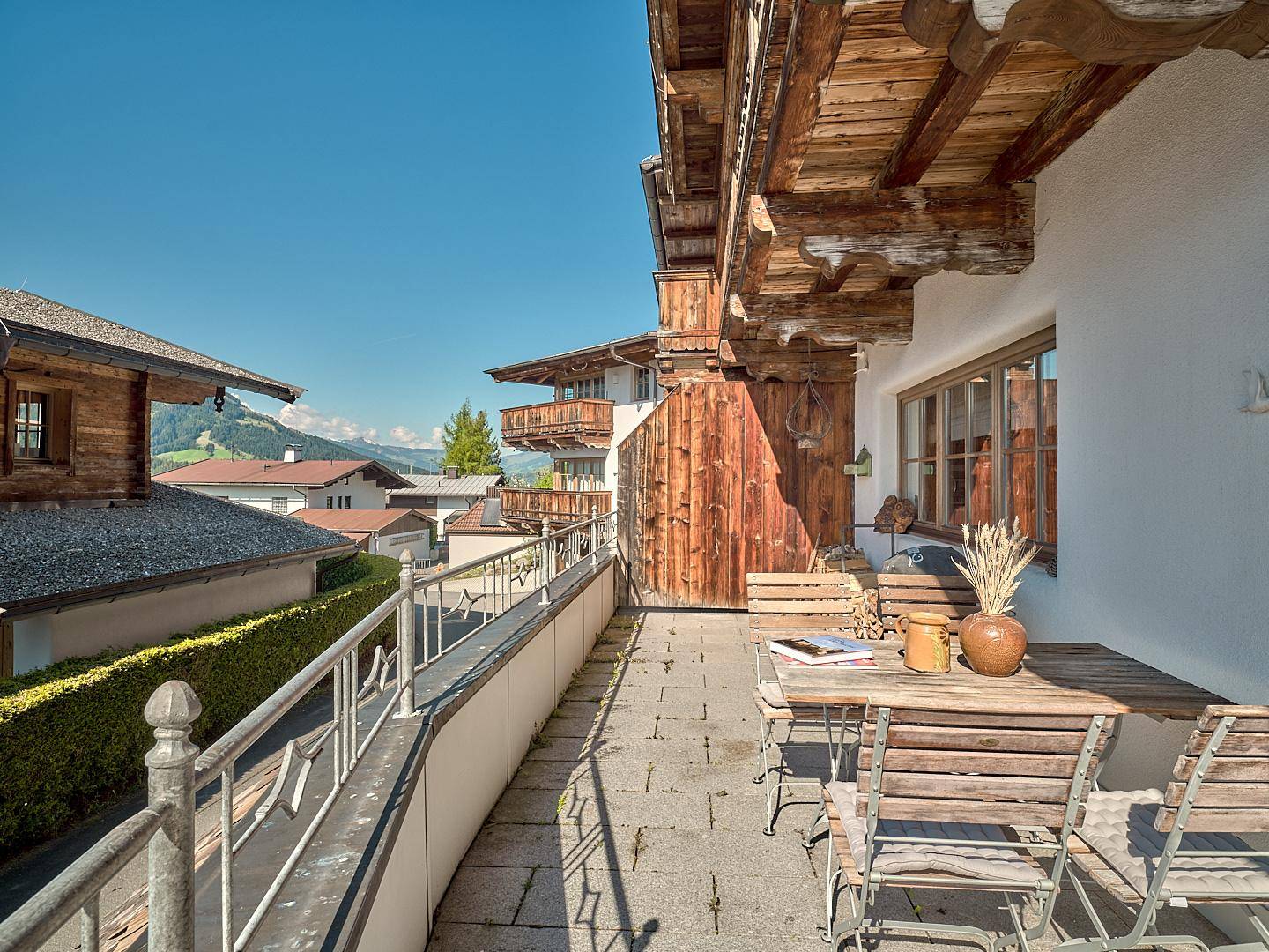 Sonniger Balkon mit Holztisch und Stühlen, umgeben von traditioneller Architektur und Bergblick.