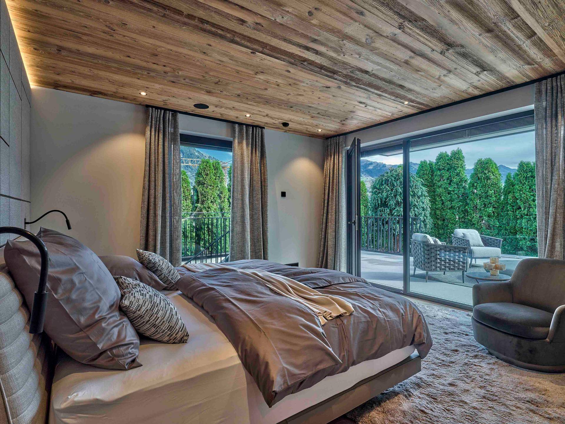 Gemütliches Schlafzimmer mit Holzbalkendecke, großen Glasfronten und Zugang zu einem Balkon mit Bergblick.