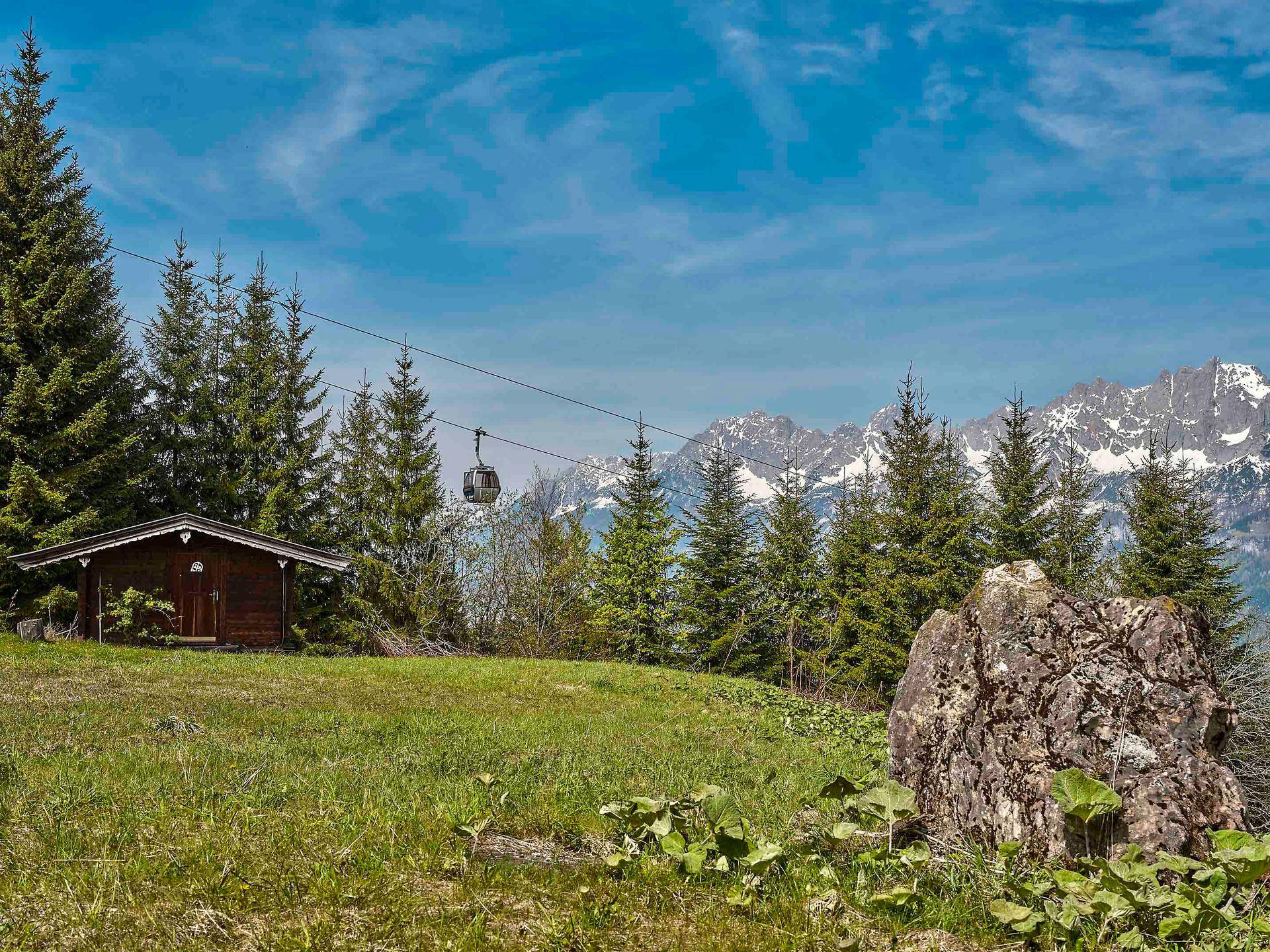 Idyllische Berglandschaft mit einer kleinen Holzhütte, Nadelbäumen und schneebedeckten Gipfeln.