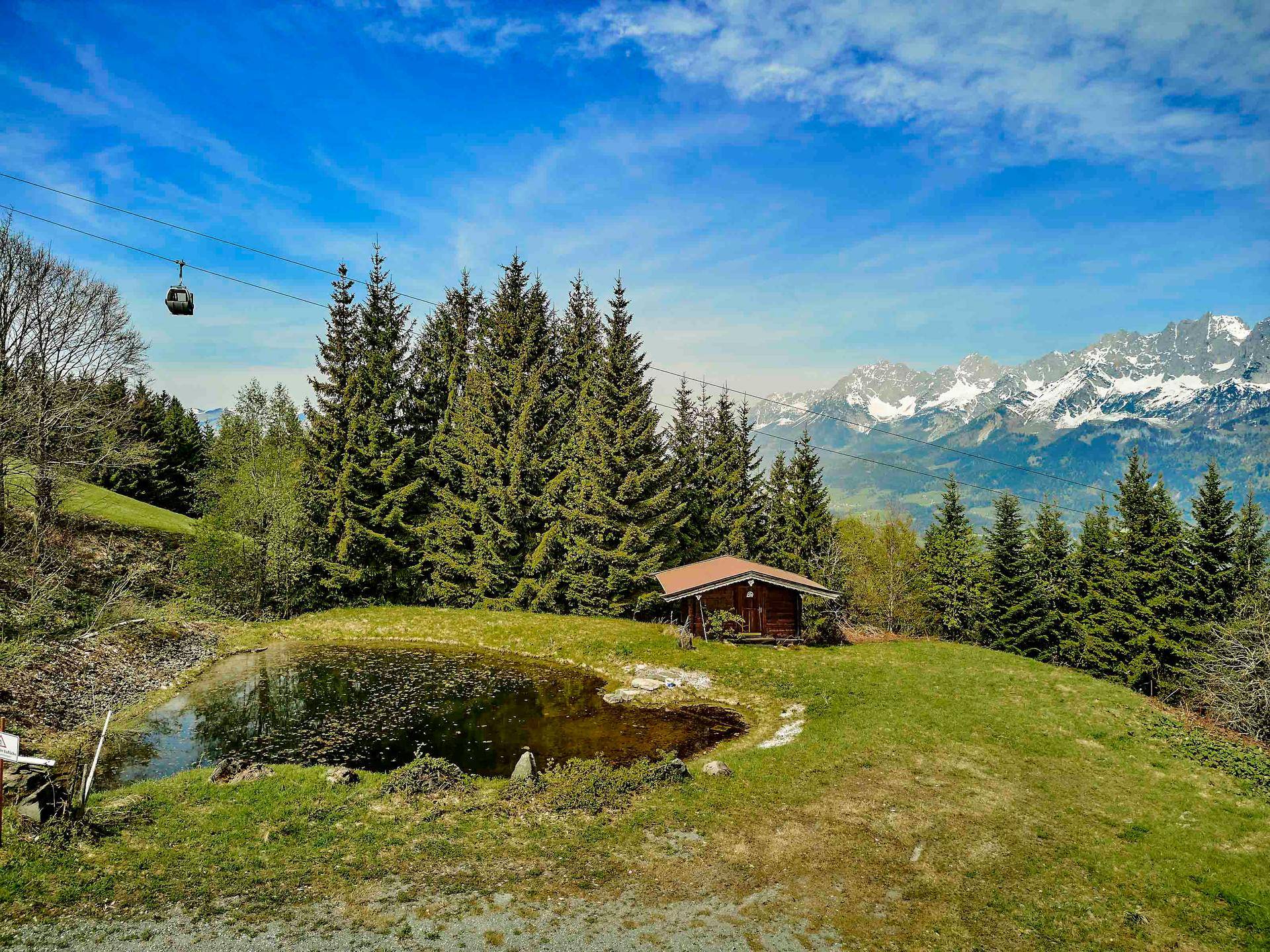 Malerische Bergkulisse mit einem kleinen Teich, einer Holzhütte und einer Seilbahn im Hintergrund.