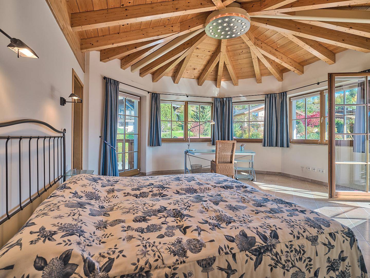 Gemütliches Schlafzimmer mit floralem Bettbezug, Holzbalkendecke und vielen Fenstern mit Grünblick.