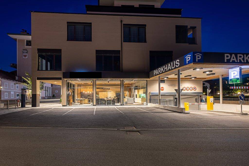 Gebäudeansicht bei Nacht mit Parkhaus und beleuchteten Geschäftsräumen.