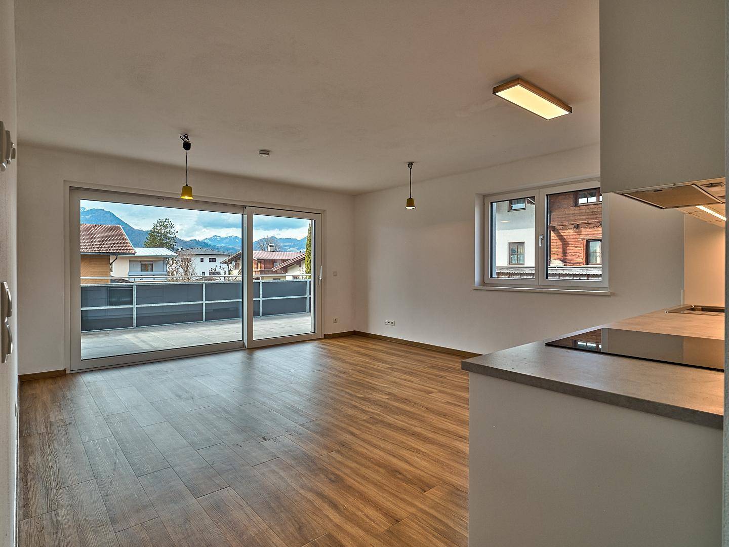 Heller, leerer Raum mit Küchenzeile, Laminatboden und Zugang zu einem großen Balkon mit Bergblick.