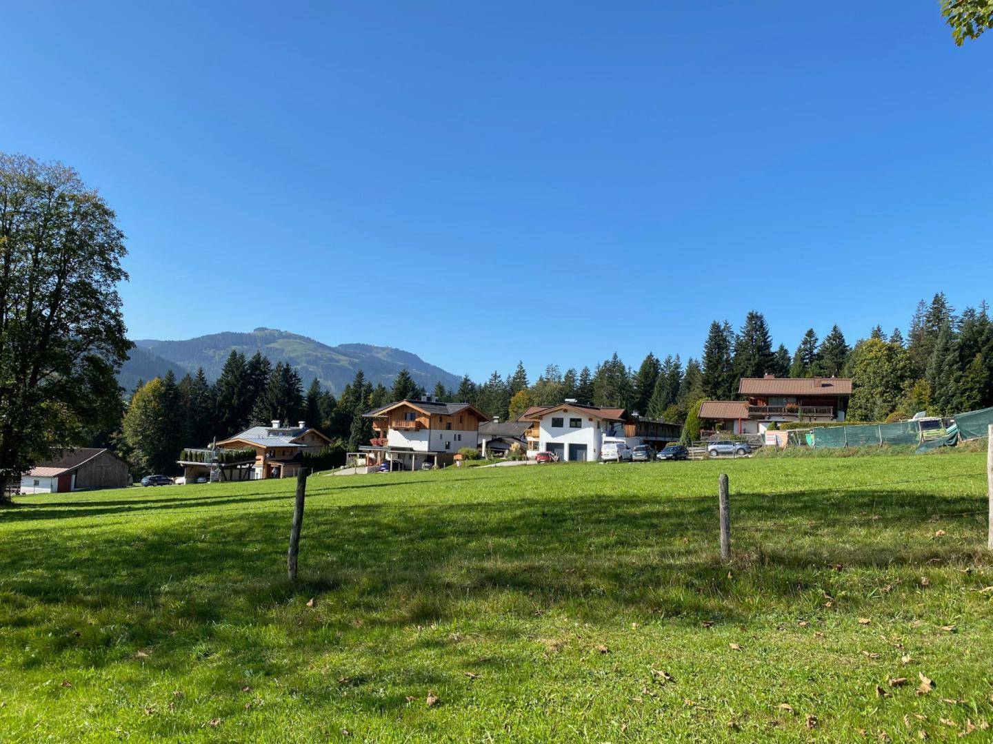 Mehrere Häuser in einer ländlichen Umgebung mit grünen Wiesen und bewaldeten Bergen im Hintergrund.