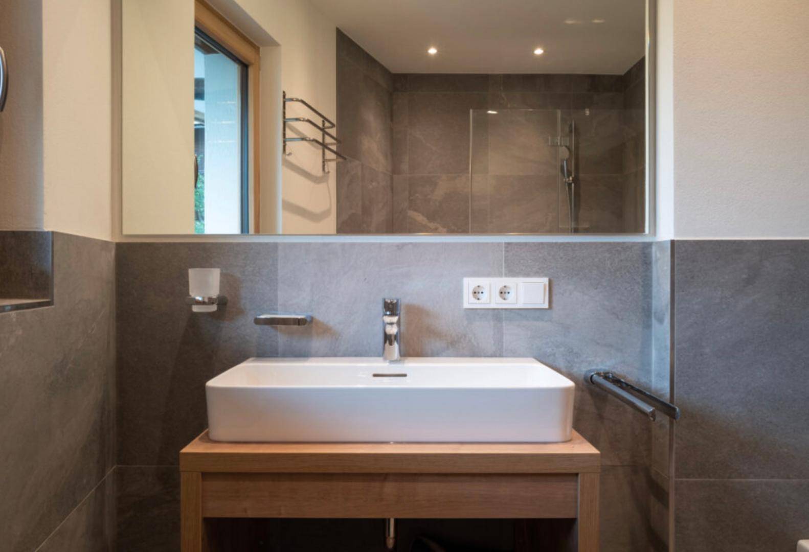 Modernes Badezimmer mit Einzelwaschbecken, großem Spiegel und grauen Wandfliesen.