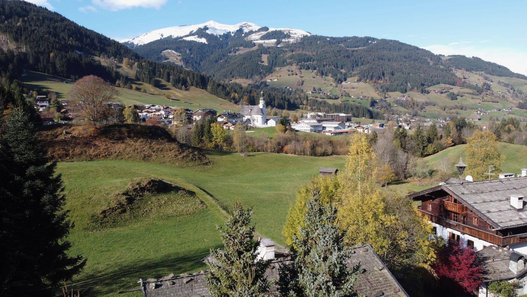Malerischer Blick auf ein alpines Dorf mit Kirche, umgeben von grünen Hügeln und schneebedeckten Bergen.