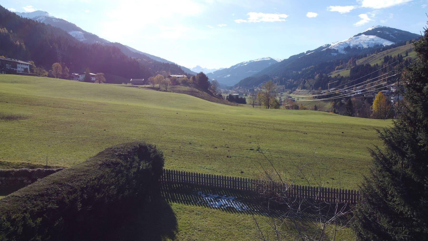 Sonniger Ausblick auf eine grüne Tallandschaft mit Bergen und einem Dorf, teilweise durch eine Hecke begrenzt.