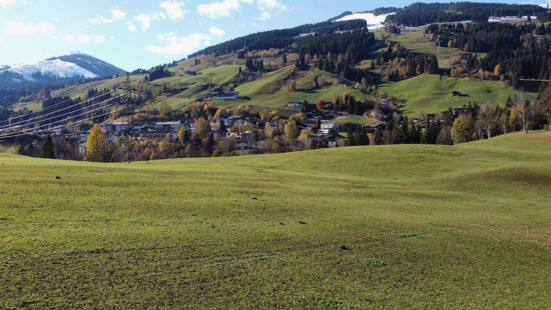 Weitläufige grüne Wiesen mit einem Dorf und bewaldeten Hängen, sowie schneebedeckten Bergen im Hintergrund.