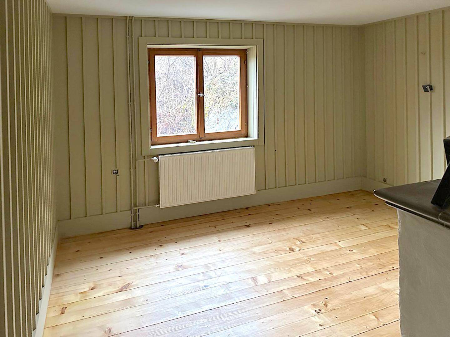 Leerer Raum mit hellgrünen Holzpaneelen, Dielenboden und einem Fenster mit Heizkörper.