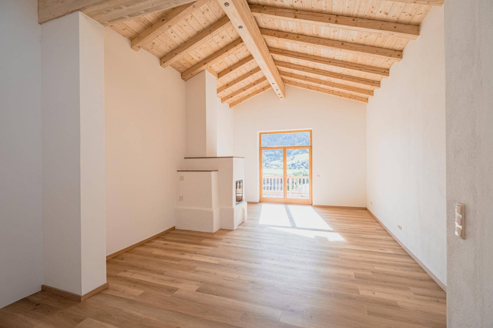 Heller Wohnbereich mit Holzbalkendecke, Kamin und Zugang zu einem Balkon mit Bergblick.
