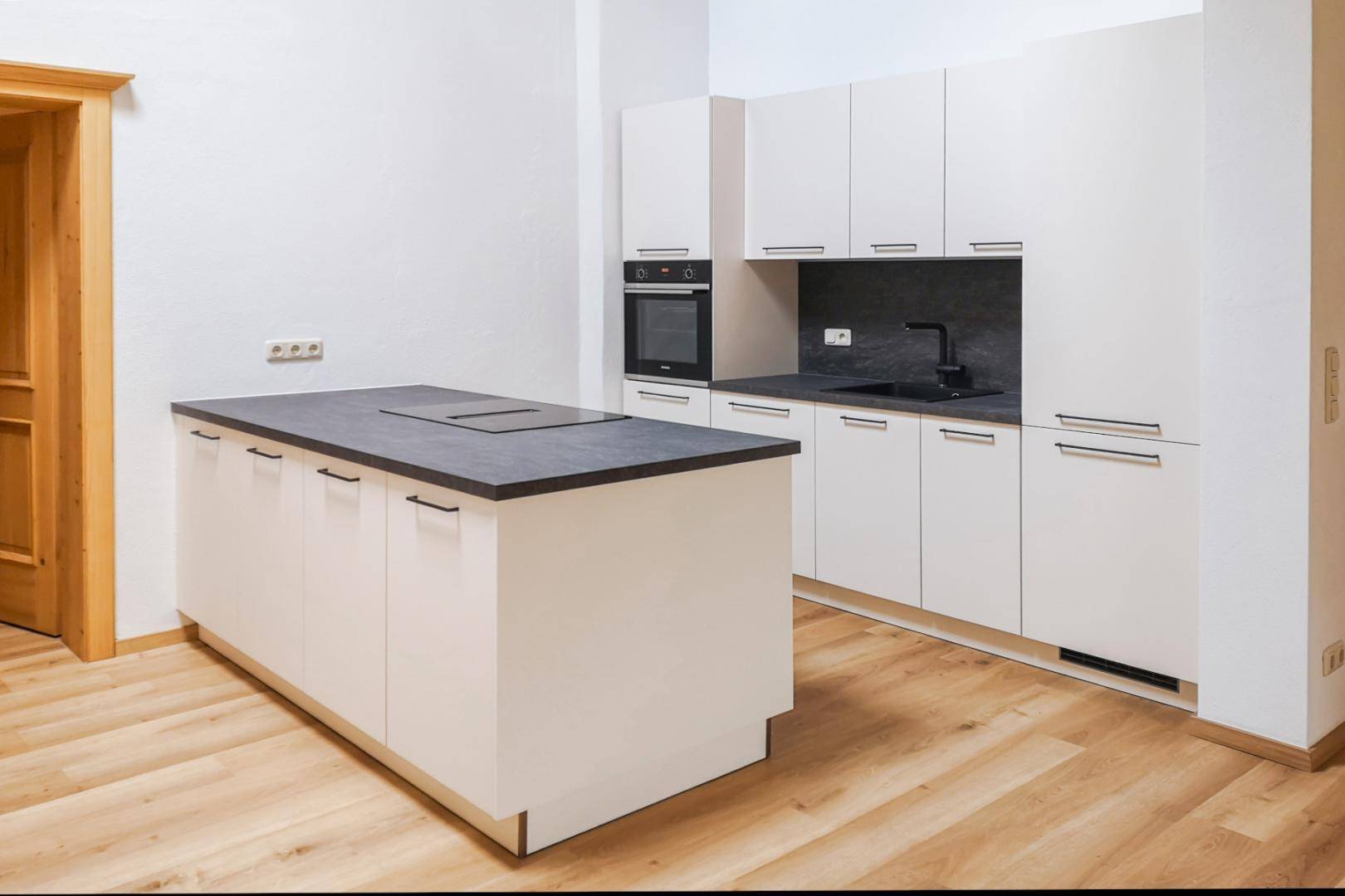Moderne Einbauküche mit weißen Schränken, dunkler Arbeitsplatte und Kochinsel mit Induktionskochfeld.