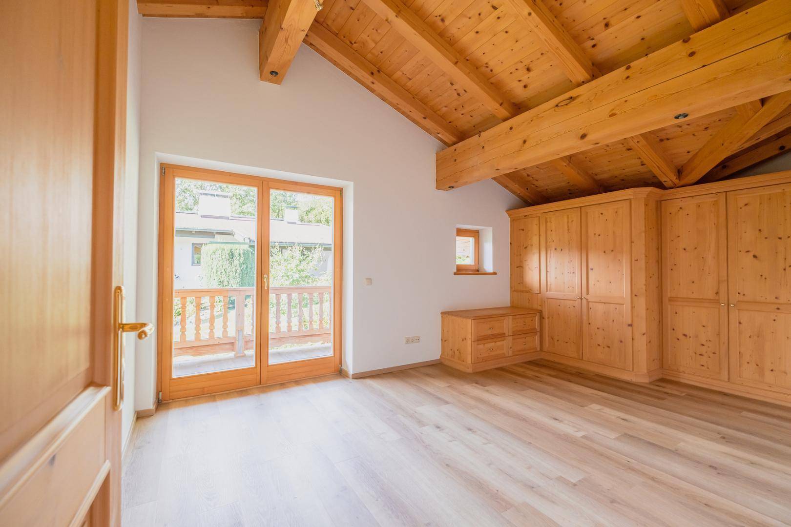Schlafzimmer mit Holzbalkendecke, Einbauschränken aus Holz und Balkonzugang mit Grünblick.