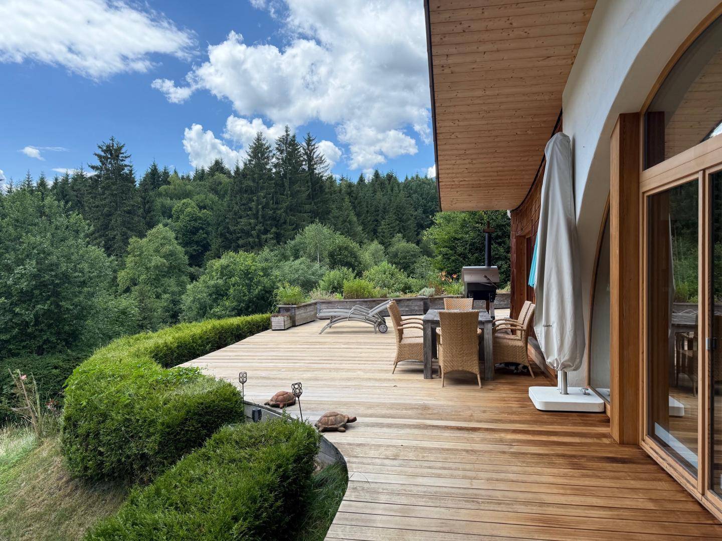 Große Holzterrasse mit Essbereich und Liegestühlen, umgeben von üppigem Grün.