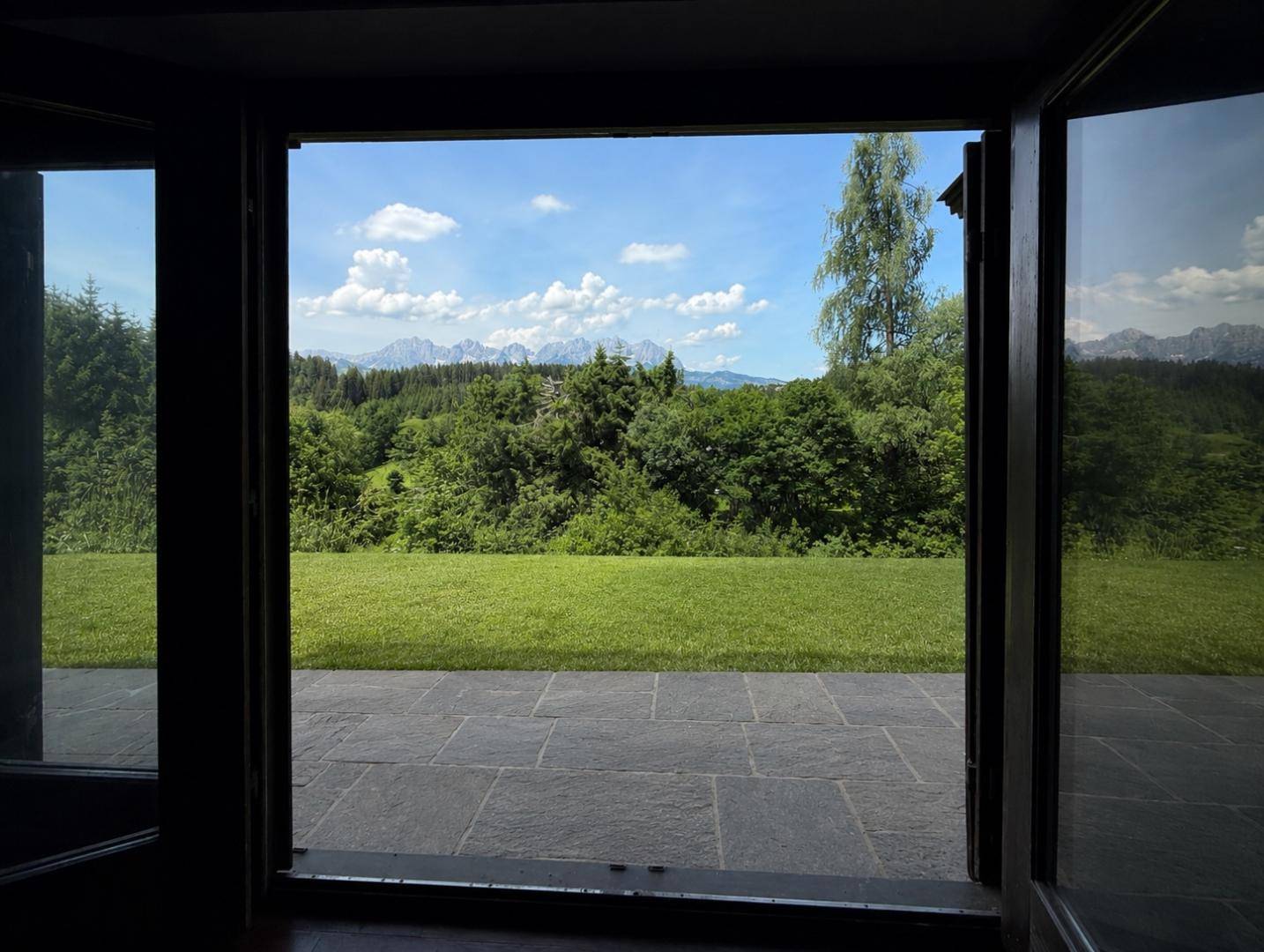 Blick von innen durch eine offene Tür auf eine Steinterrasse und grüne Landschaft.
