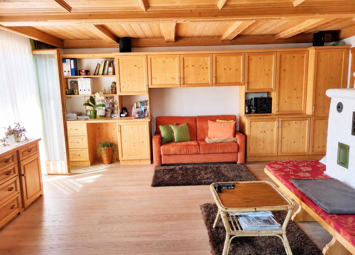 Helles Wohnzimmer mit traditionellem Kachelofen, Holzbalkendecke und integriertem Arbeitsbereich.