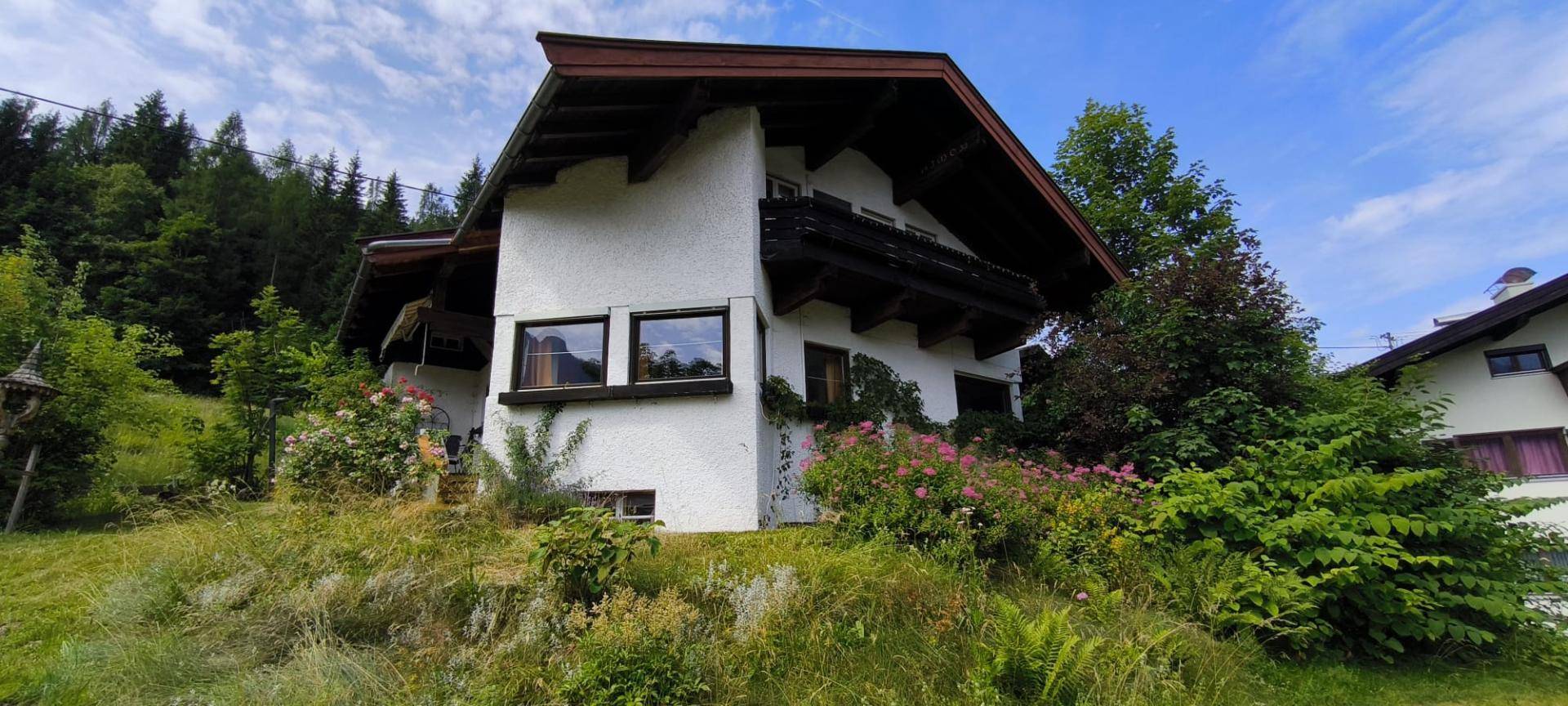 Alpines Haus mit Holzbalkon und blühenden Sträuchern in einer grünen Hügellandschaft.