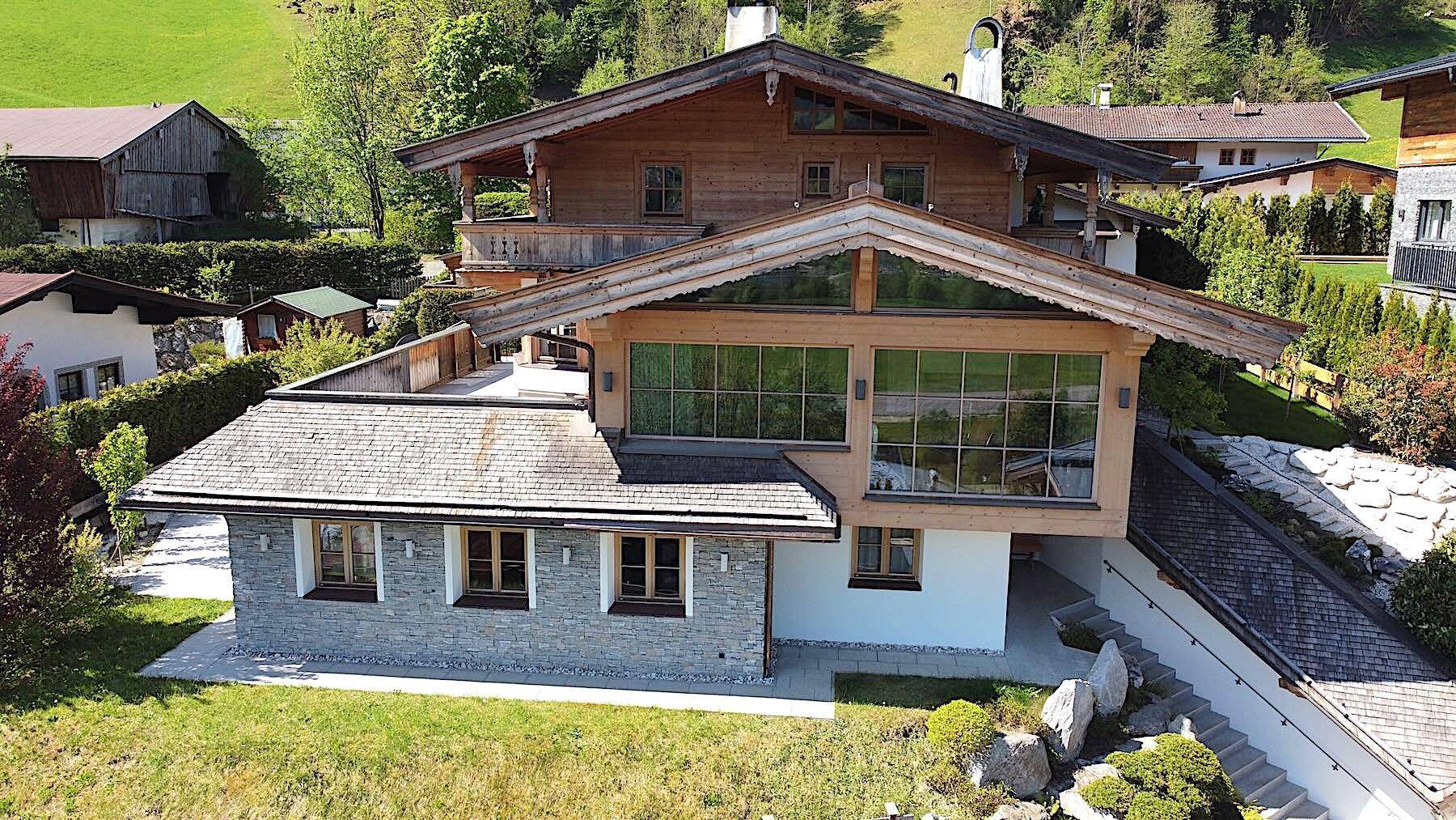 Ansicht des Hauses mit traditioneller Holzfassade und großen Fensterfronten, umgeben von grünen Hügeln.