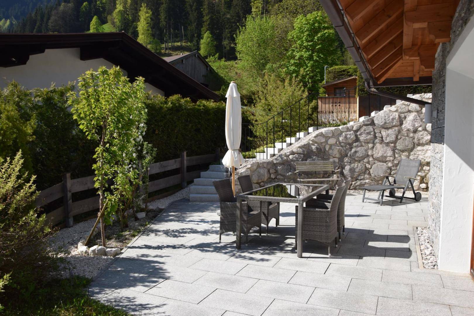 Gepflasterte Terrasse mit Gartenmöbeln und Sonnenschirm, umgeben von grüner Bepflanzung.