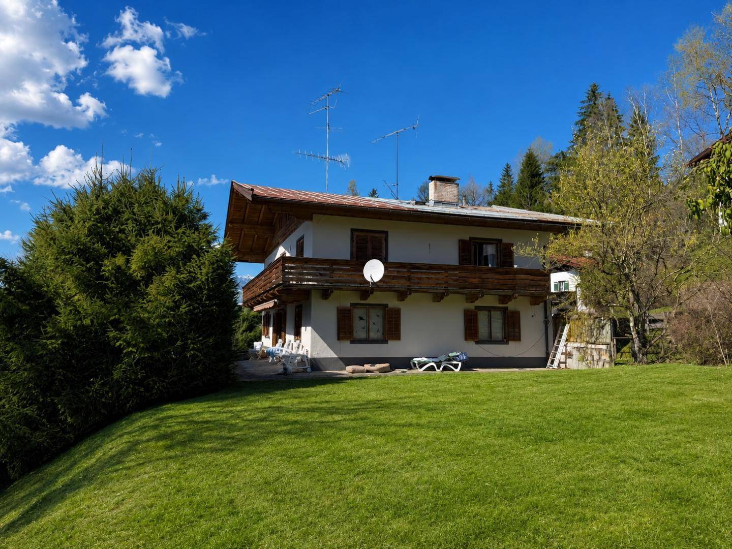 Freistehendes Haus mit großem Rasen und Holzbalkon in sonniger alpiner Landschaft.