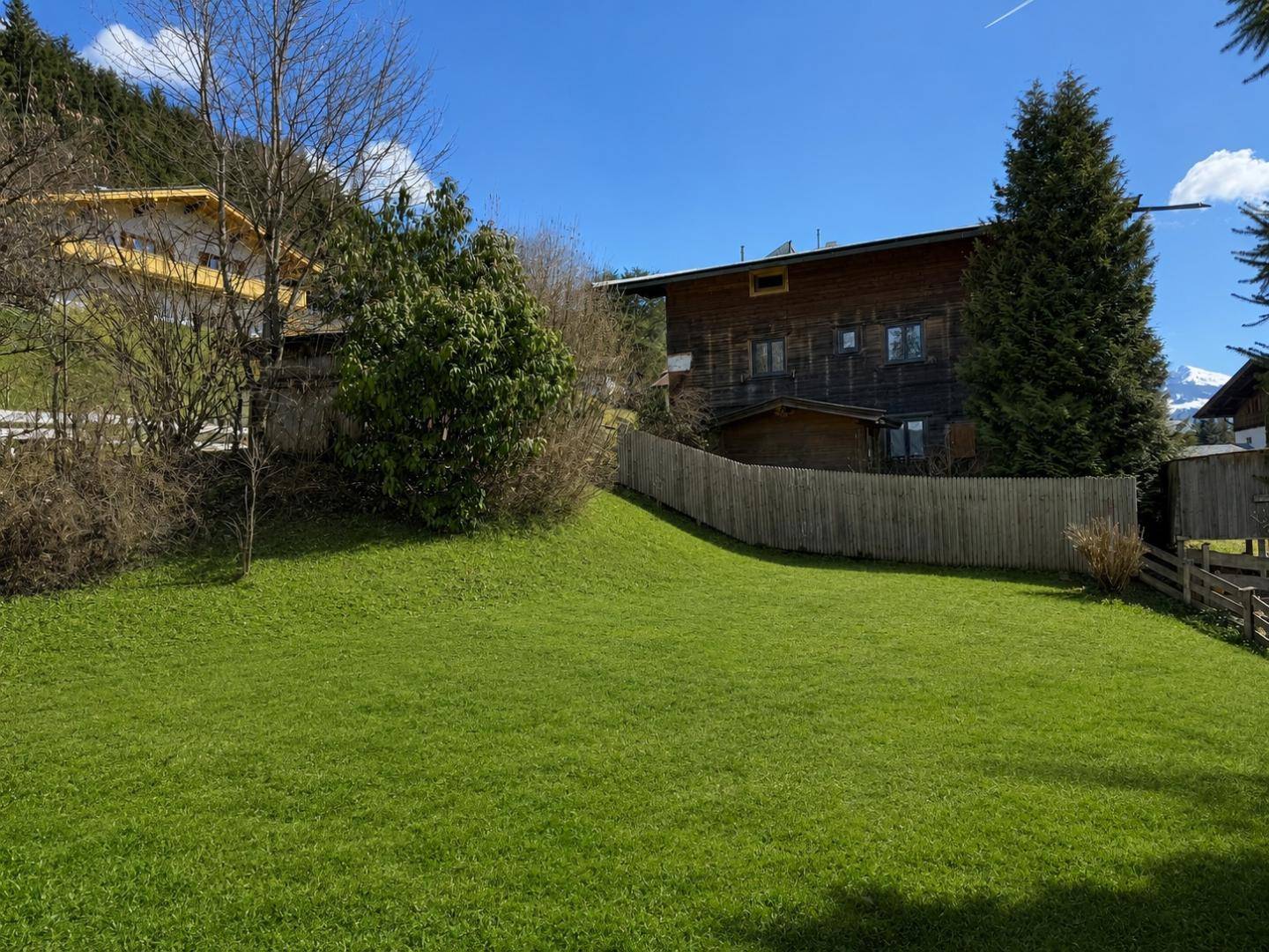Großzügiger grüner Rasen mit Holzzaun und einem traditionellen Holzhaus im Hintergrund.