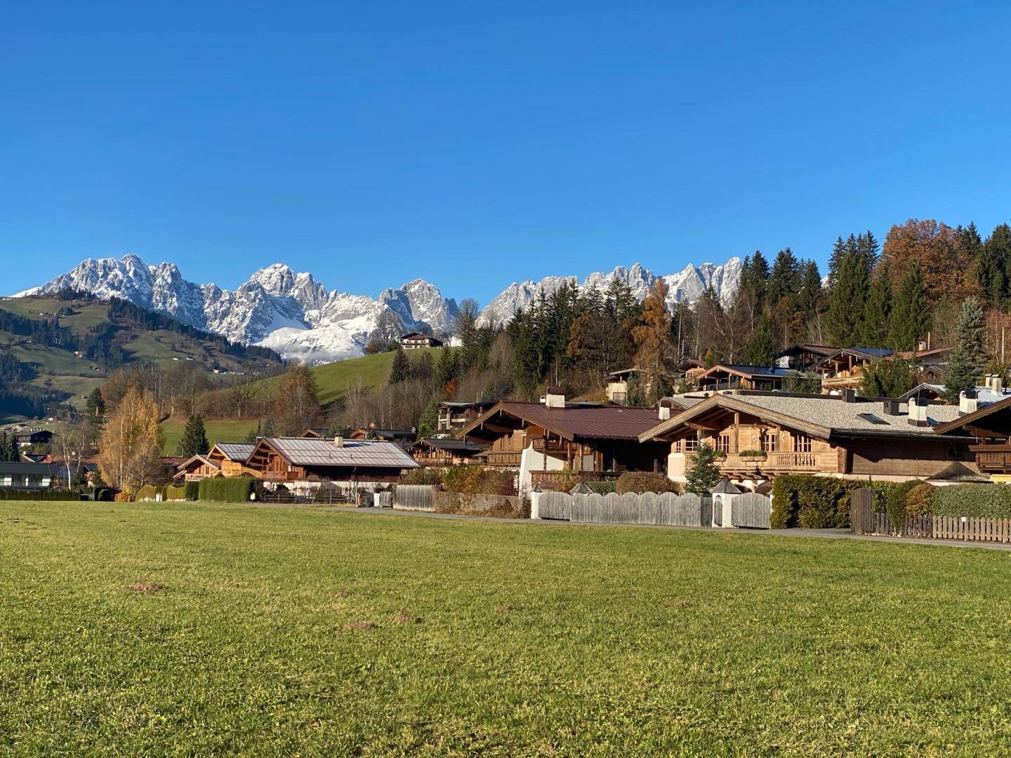 Blick über ein grünes Feld auf traditionelle Häuser und schneebedeckte Berge in der Ferne.