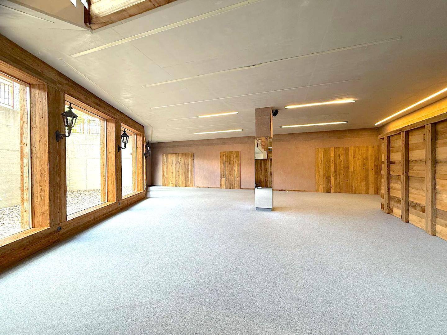 Großer, heller Raum mit Teppichboden, Holzwänden und einer Fensterfront mit Blick nach draußen, sehr einladend.