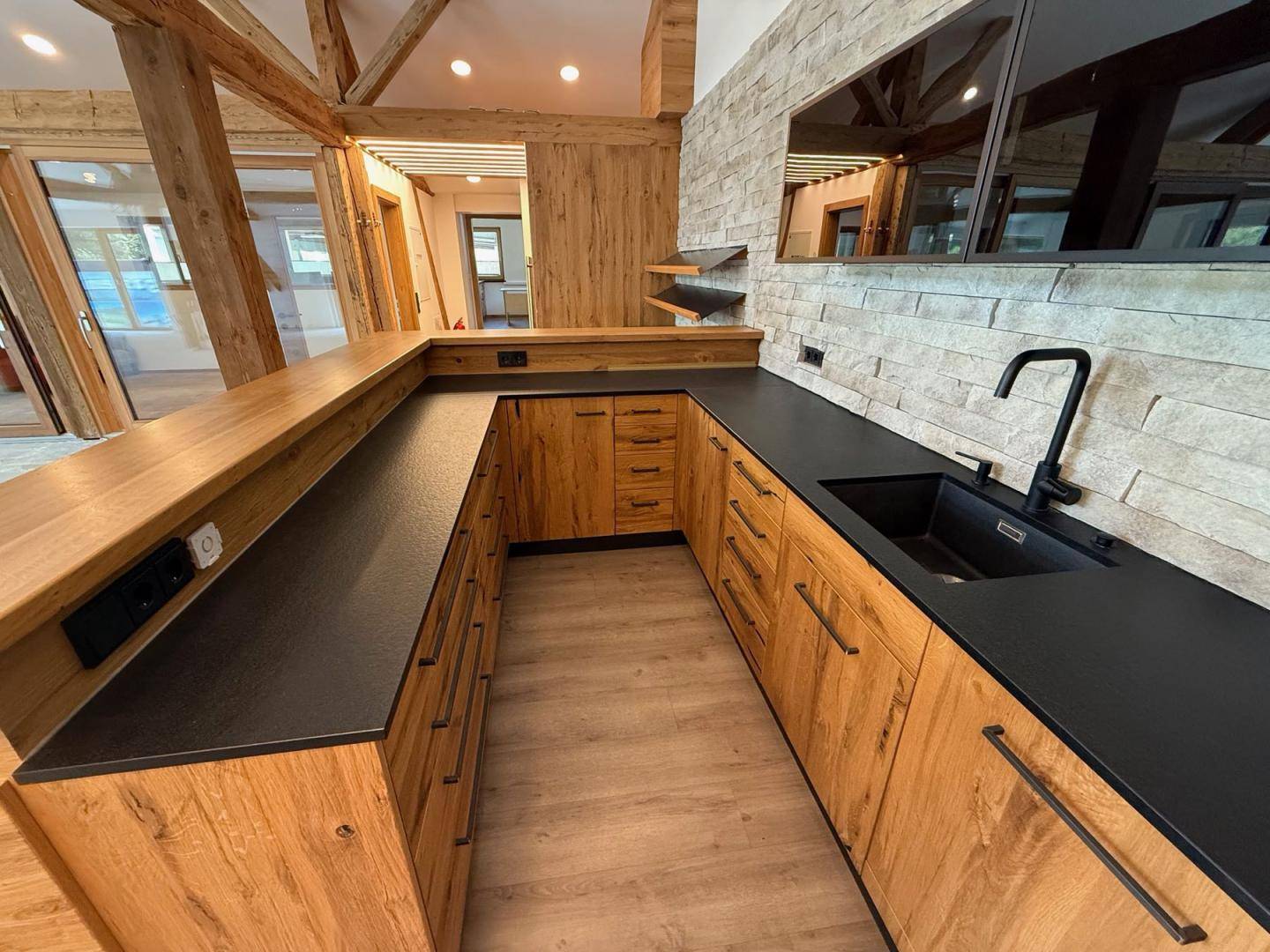 Detailansicht einer modernen Holzküche mit schwarzer Spüle und Arbeitsplatte, sowie Steinwand.
