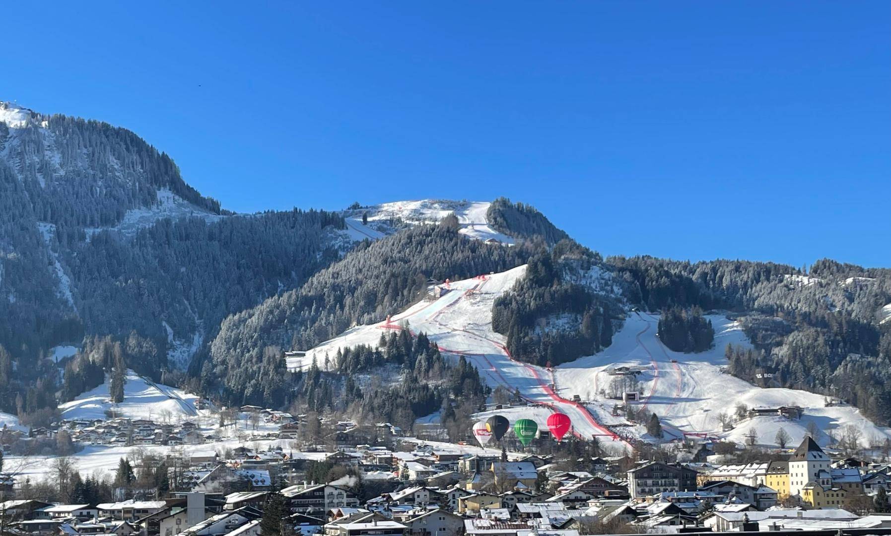 Winterliche Berglandschaft mit verschneiten Skipisten, Dorf und bunten Heißluftballons.