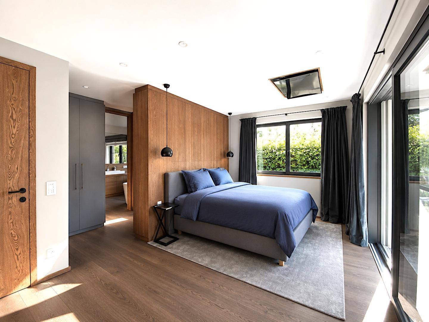 Modernes Schlafzimmer mit Holzakzentwand, großen Fenstern und Zugang zu einem Bad en Suite.
