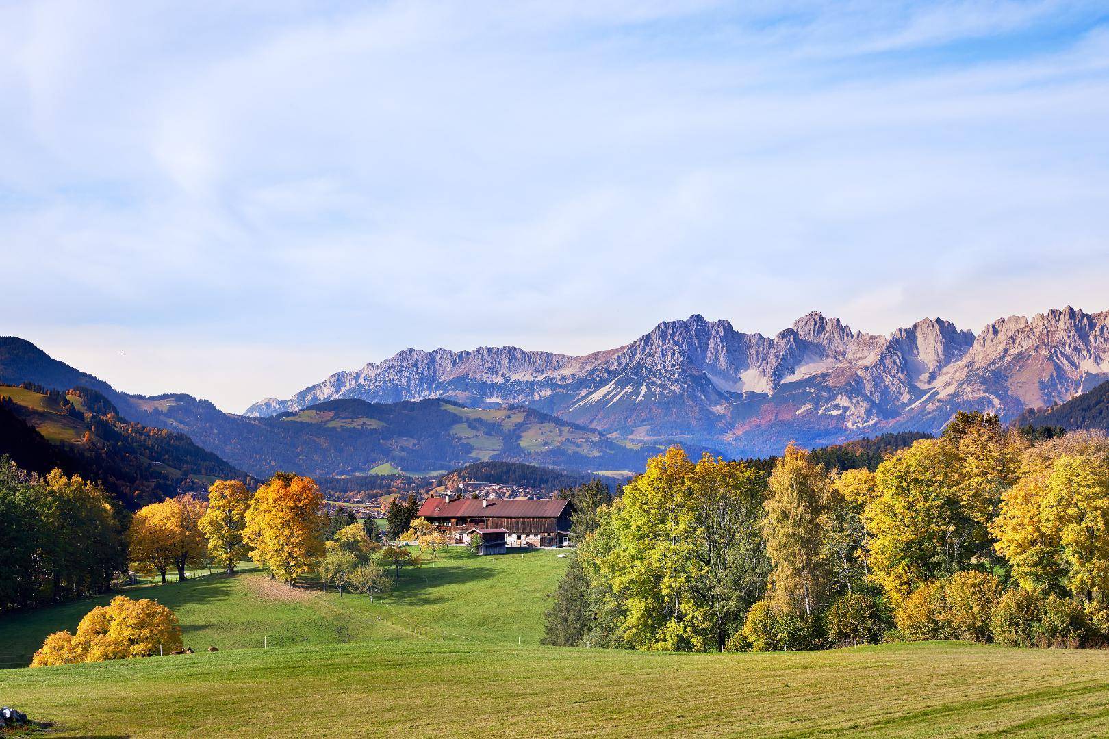 Malerische Berglandschaft mit grünen Wiesen, herbstlichen Bäumen und einem Haus in der Ferne.