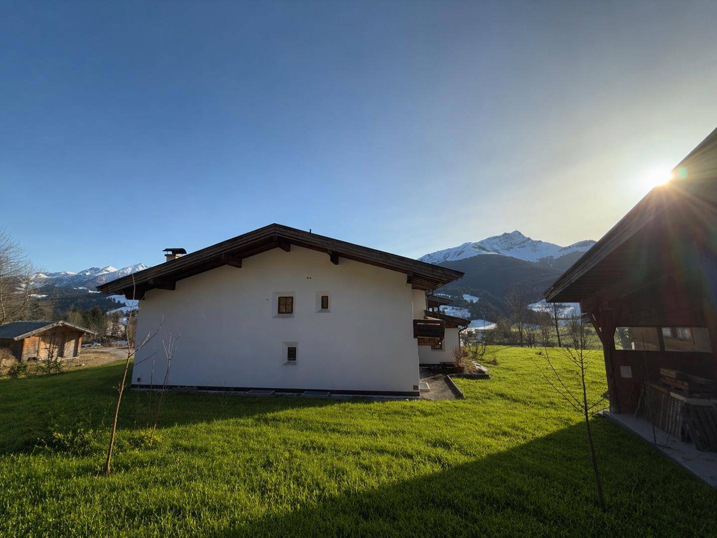 Seitenansicht eines Hauses mit weißer Fassade und dunklem Dach vor einer Kulisse schneebedeckter Berge.
