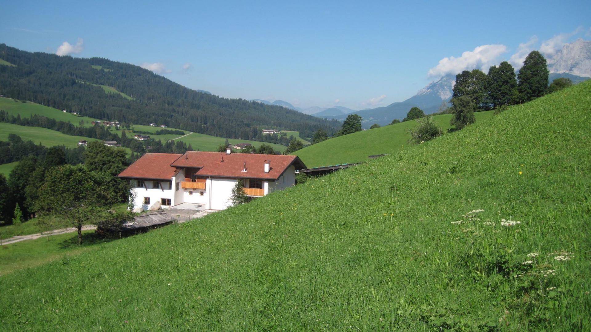 Traditionelles Haus mit rotem Dach und weißer Fassade, eingebettet in eine grüne Alpenlandschaft.