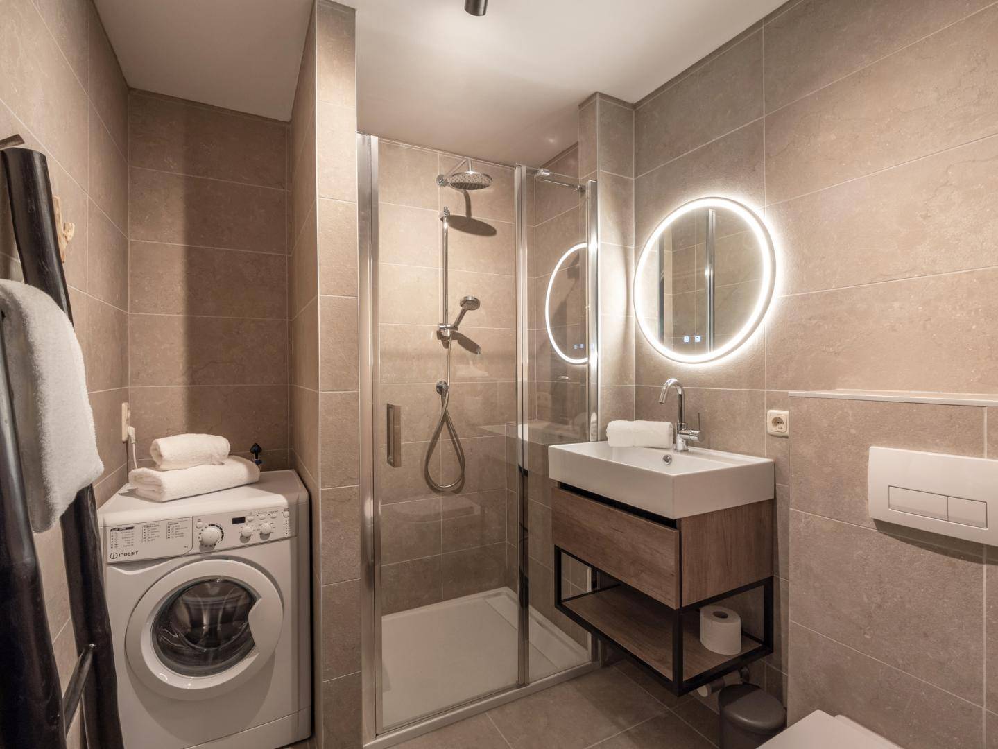 Modernes Badezimmer mit begehbarer Dusche, Waschbecken und Waschmaschine, in hellen Fliesen gehalten.