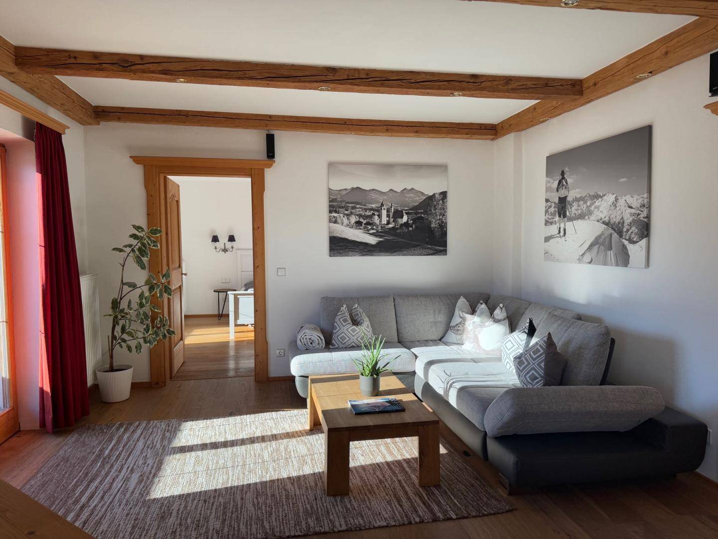 Helles Wohnzimmer mit Ecksofa, Holzbalken und Blick in den angrenzenden Raum.