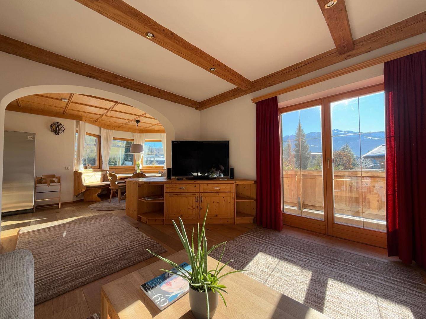 Lichtdurchfluteter Wohnbereich mit Holzbalken, TV-Möbel und Zugang zum Balkon mit Bergblick.