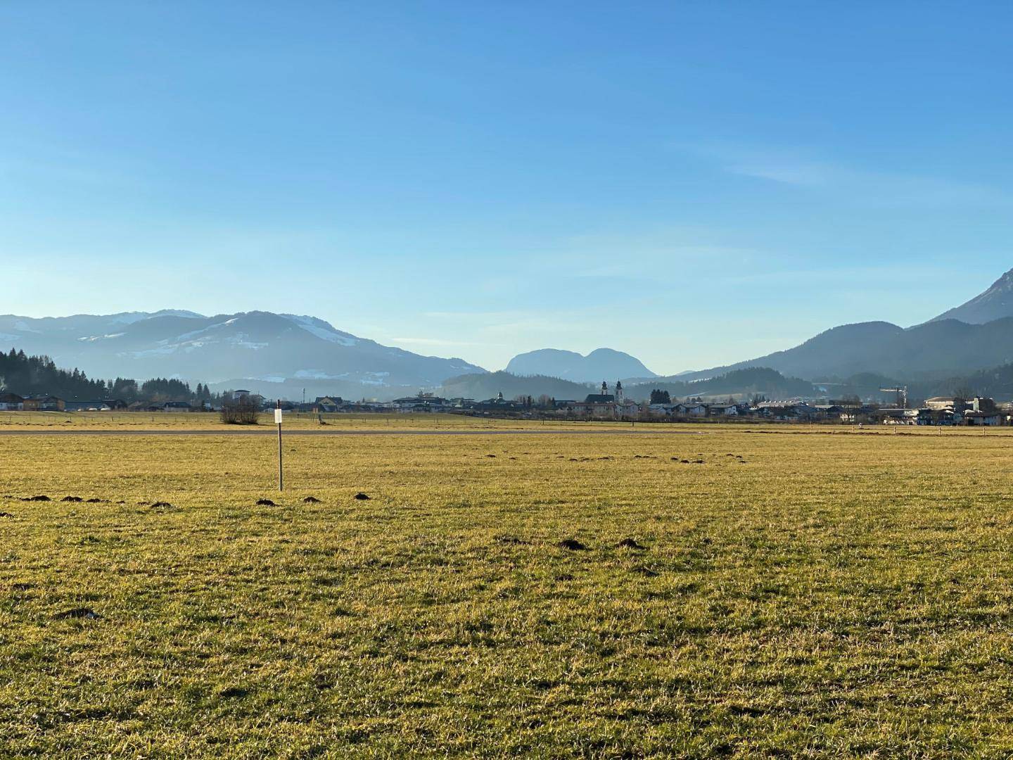 Ausgedehntes grünes Feld mit einem Dorf und sanften Hügeln in der Ferne unter blauem Himmel.