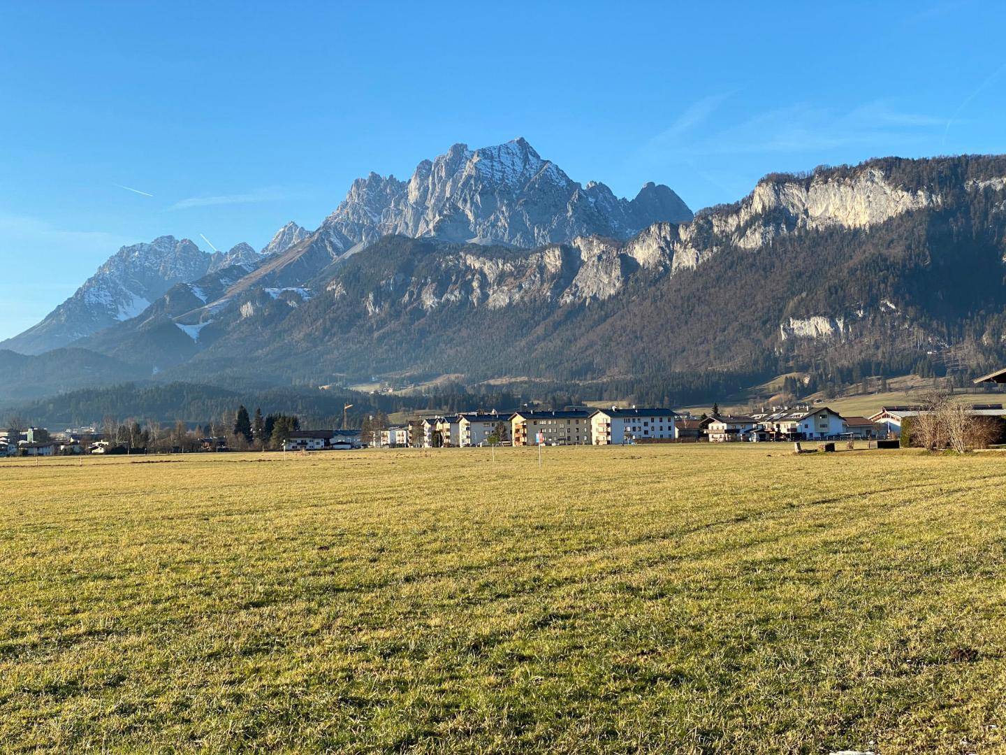 Breites grünes Feld mit einem Dorf und imposanten Bergen im Hintergrund unter strahlendem Sonnenschein.