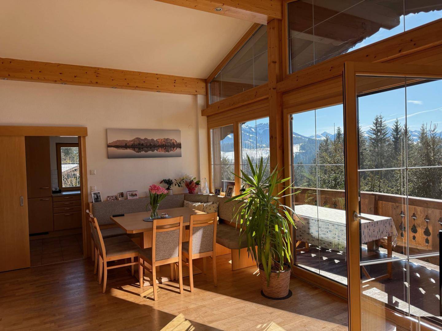 Heller Wohn- und Essbereich mit Holzbalken, großen Fenstern und Zugang zu einem Balkon mit Bergblick.