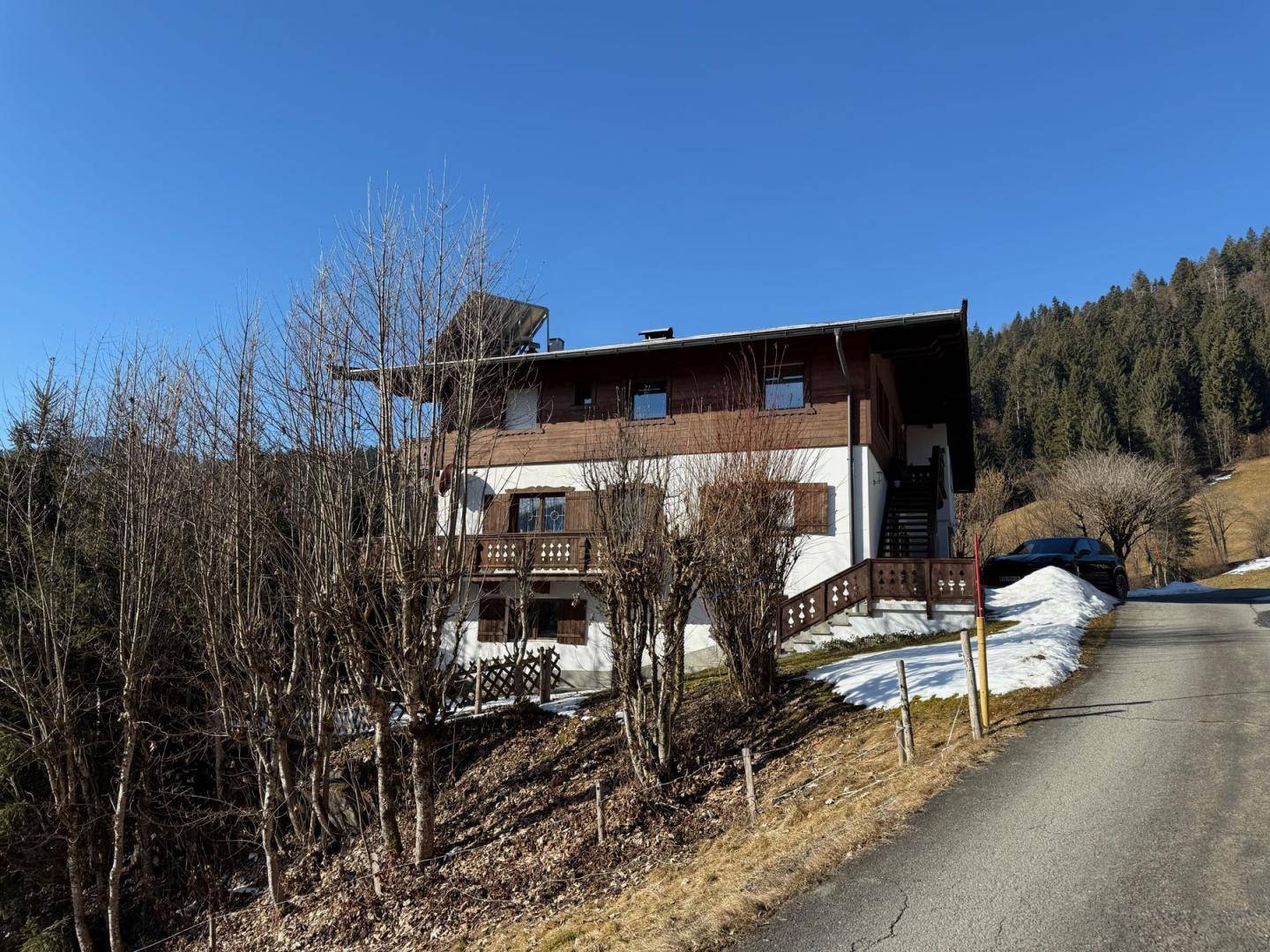 Alpenhaus im Winter mit schneebedecktem Boden, kahlen Bäumen und traditioneller Holzfassade unter blauem Himmel.