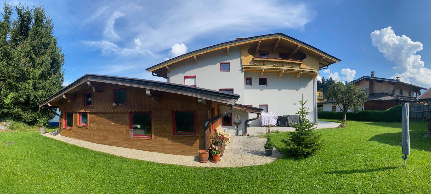 Haus mit großem Garten und Holzanbau, umgeben von hohen Bäumen und einer weitläufigen Wiese.