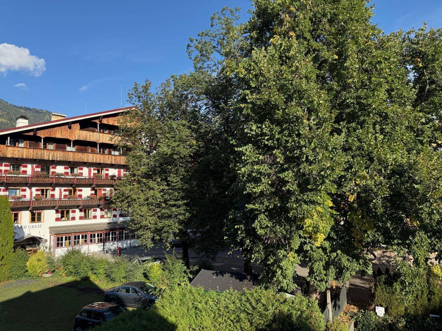 Traditionelles Gebäude mit Holzbalkonen und roten Fensterläden, umgeben von Bäumen.