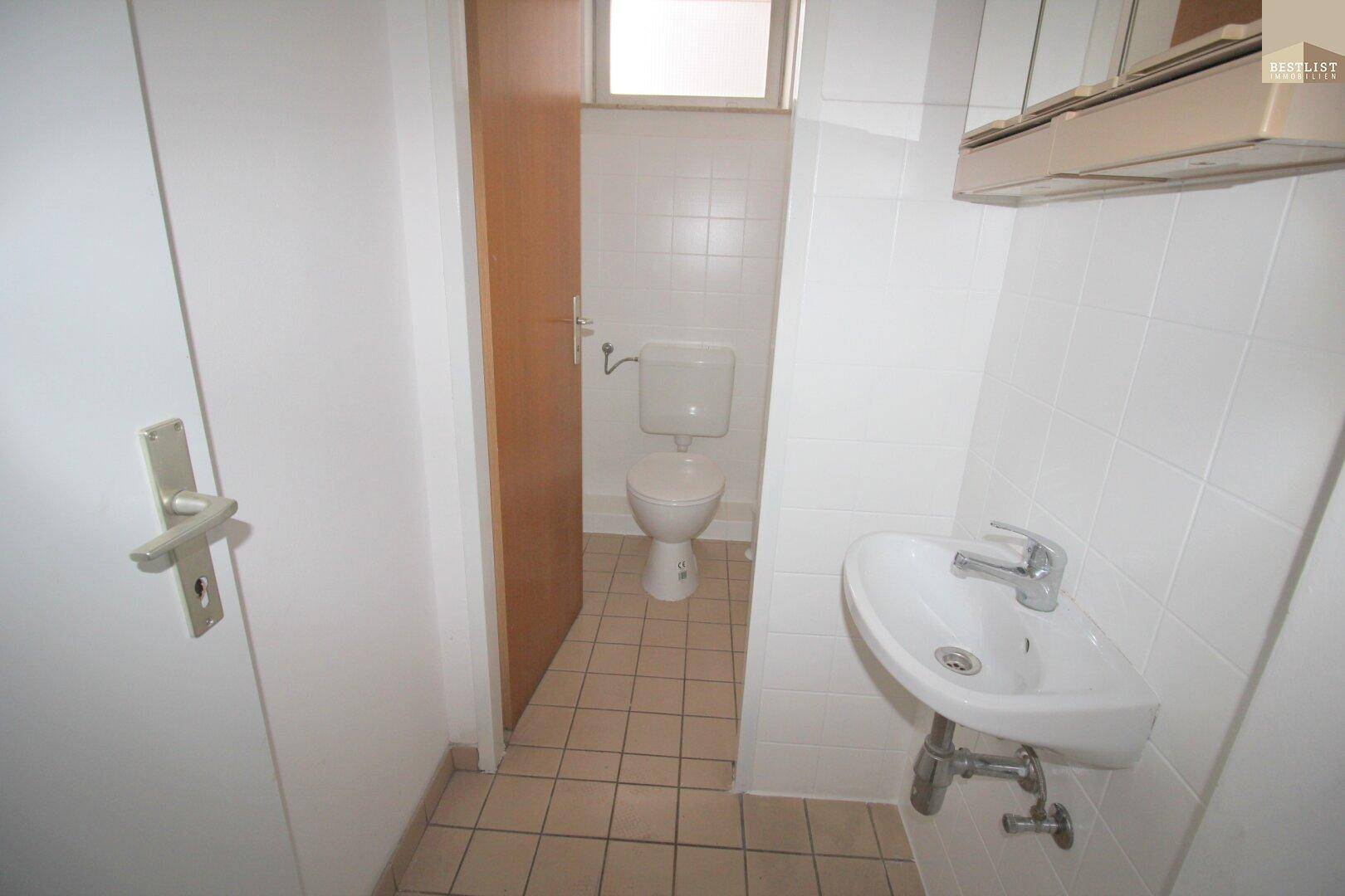 Separate Toilette mit weißen Fliesen, WC, kleinem Waschbecken und einem Fenster über der Tür.