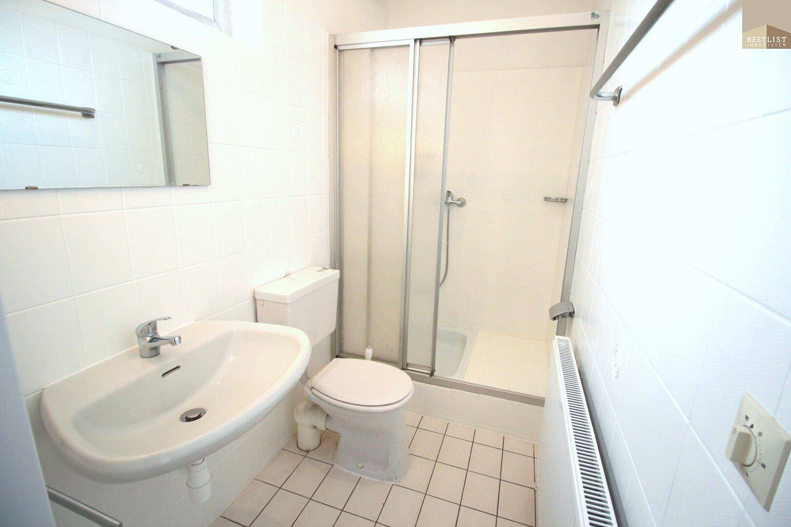 Funktionales Badezimmer mit weißen Fliesen, Waschbecken, Toilette und einer Duschkabine.
