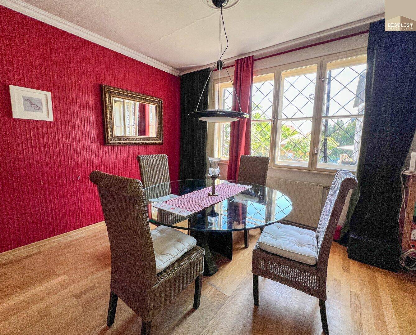 Esszimmer mit roter Wand, Holzboden und einem runden Glastisch mit Korbstühlen.