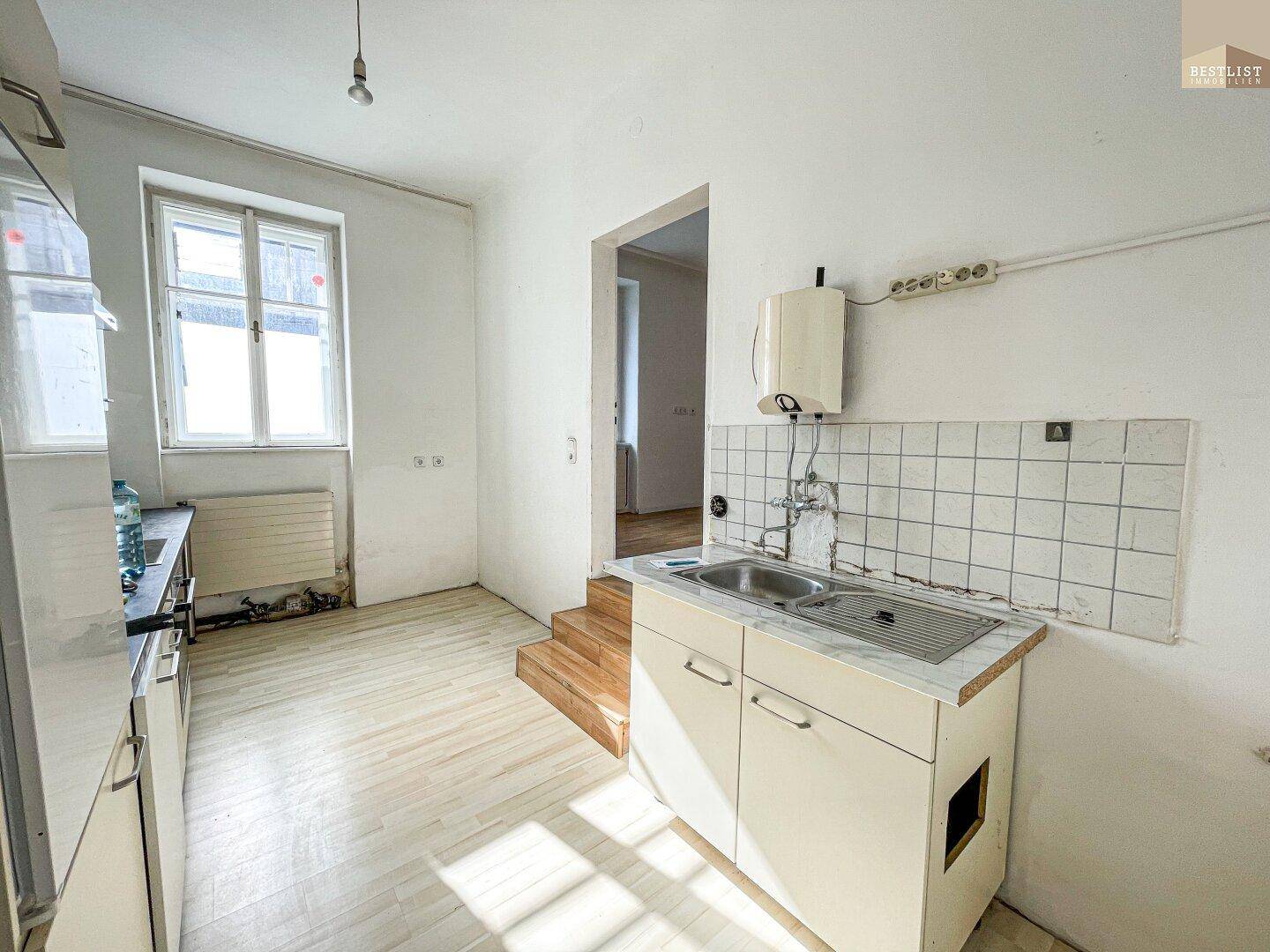 Einfache Küche mit Spülbecken, Fliesenrückwand und Fenster, in originalem Zustand und bereit für eine Modernisierung.