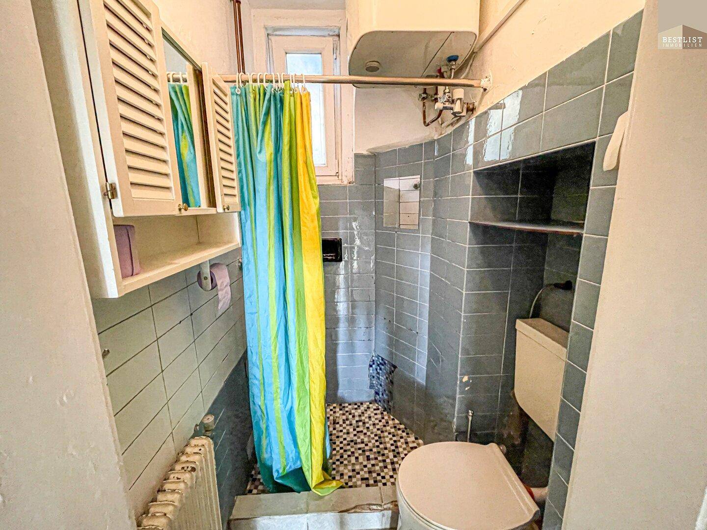 Kleines Badezimmer mit Dusche, WC und blauen Fliesen, in originalem Zustand mit Fenster.