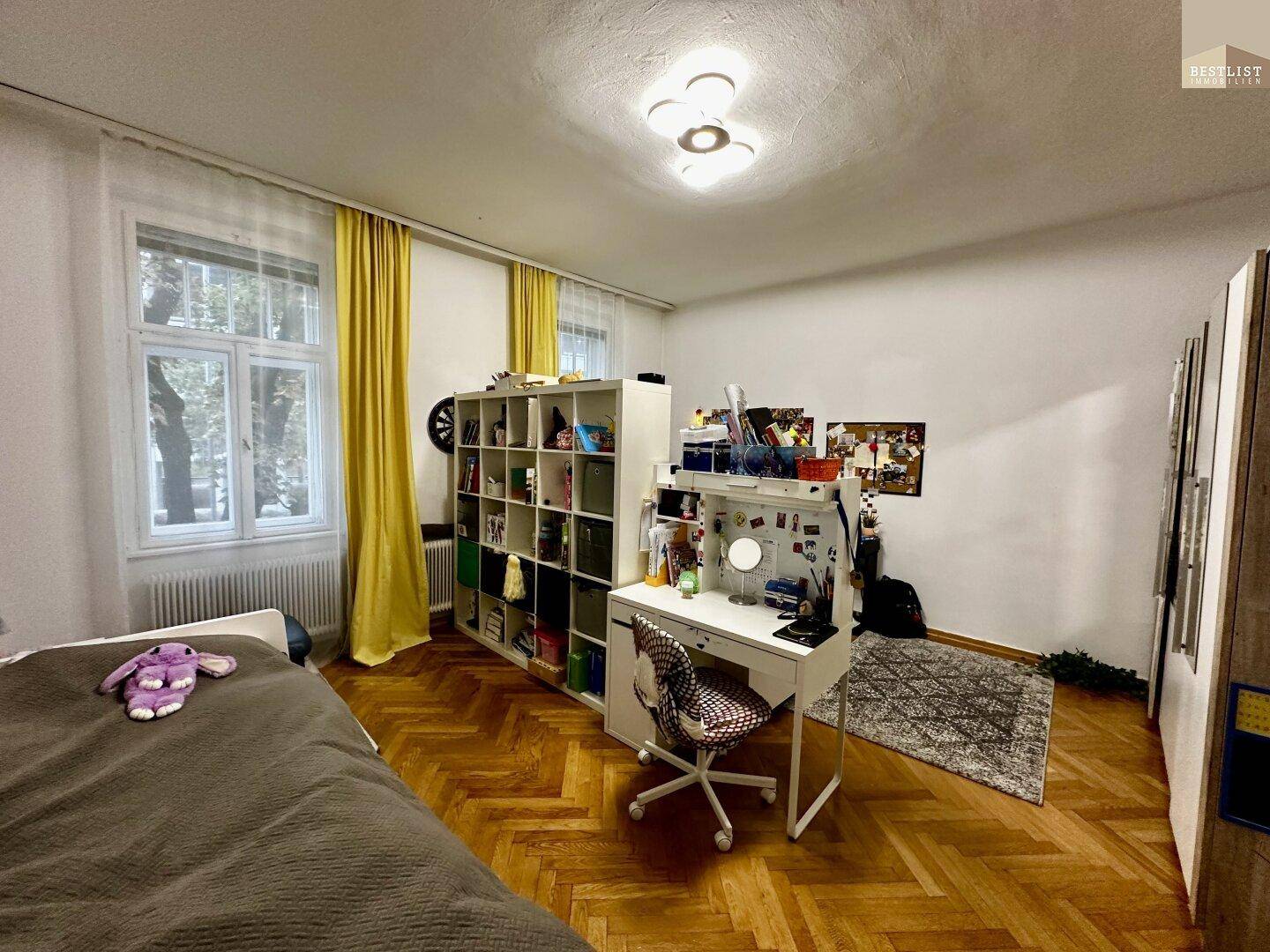 Helles Schlafzimmer mit Parkettboden, Schreibtisch und Bücherregal, gut beleuchtet durch große Fenster.