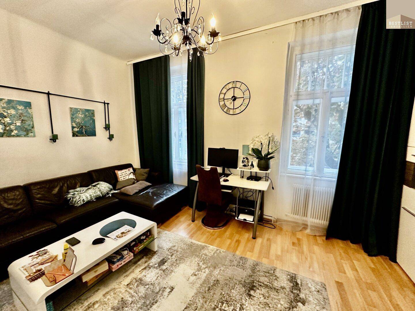 Gemütliches Wohnzimmer mit dunklem Sofa, Holzboden und einem Arbeitsbereich am Fenster mit viel Tageslicht.